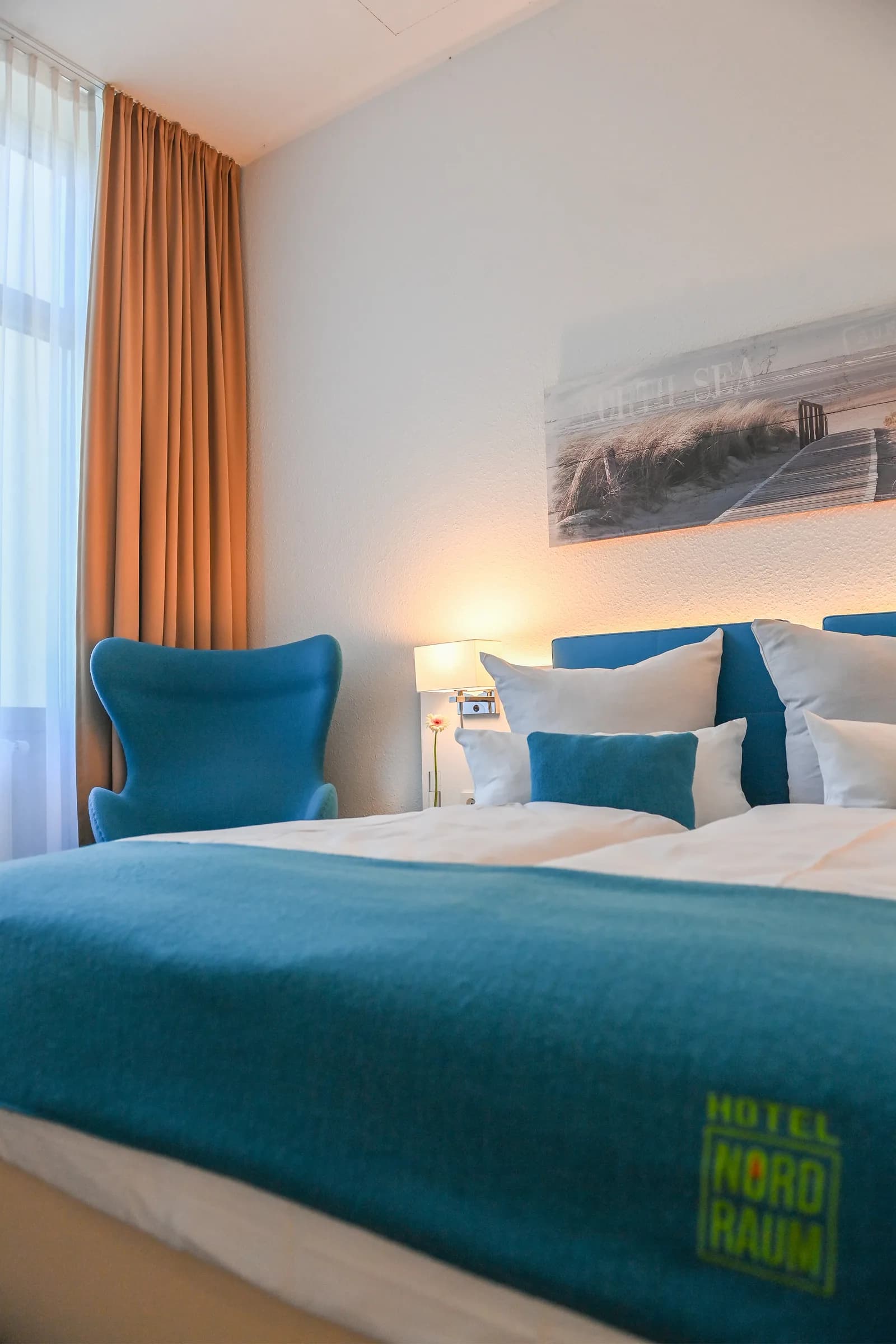 Chambre simple avec tête de lit bleue et couvre-lit orné du logo NordRaum