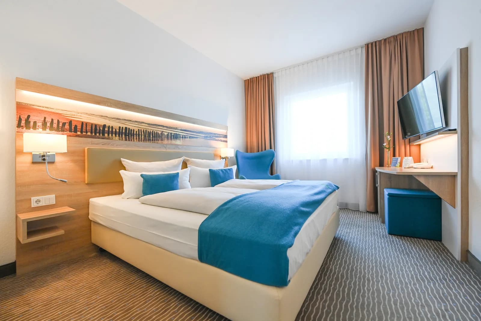Doppelzimmer mit Strandpanorama am Kopfteil, blauem Sessel und Schreibtisch